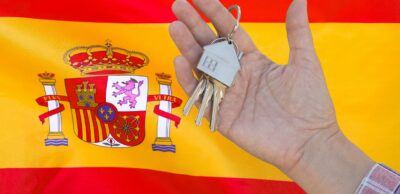 Hand holding house keys with a keyring shaped like a home, set against the Spanish flag.
